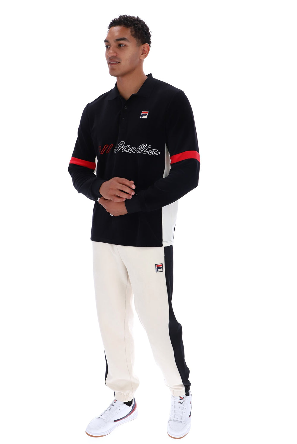 A man modeling a black and white Fila long-sleeve polo shirt with red accents on the sleeves, paired with white pants and white sneakers, against a plain white background.