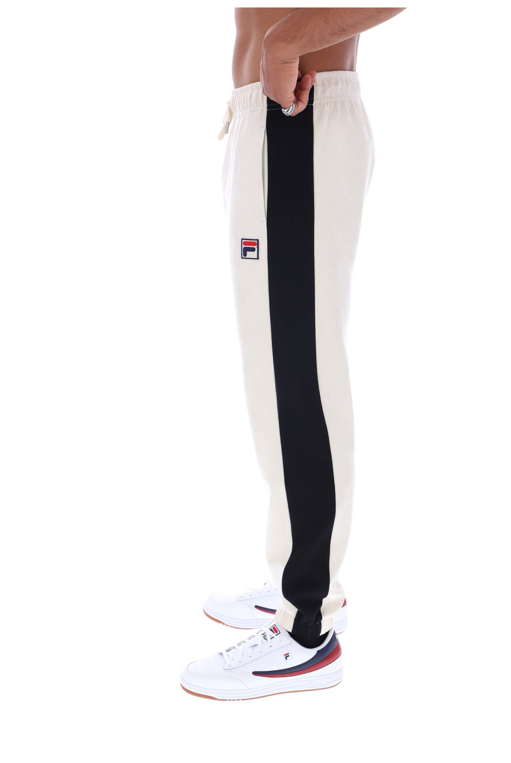 Side view of a person wearing off-white sweatpants with a bold black vertical stripe down the side and a small Fila logo patch near the upper thigh, paired with white sneakers on a plain white background.
