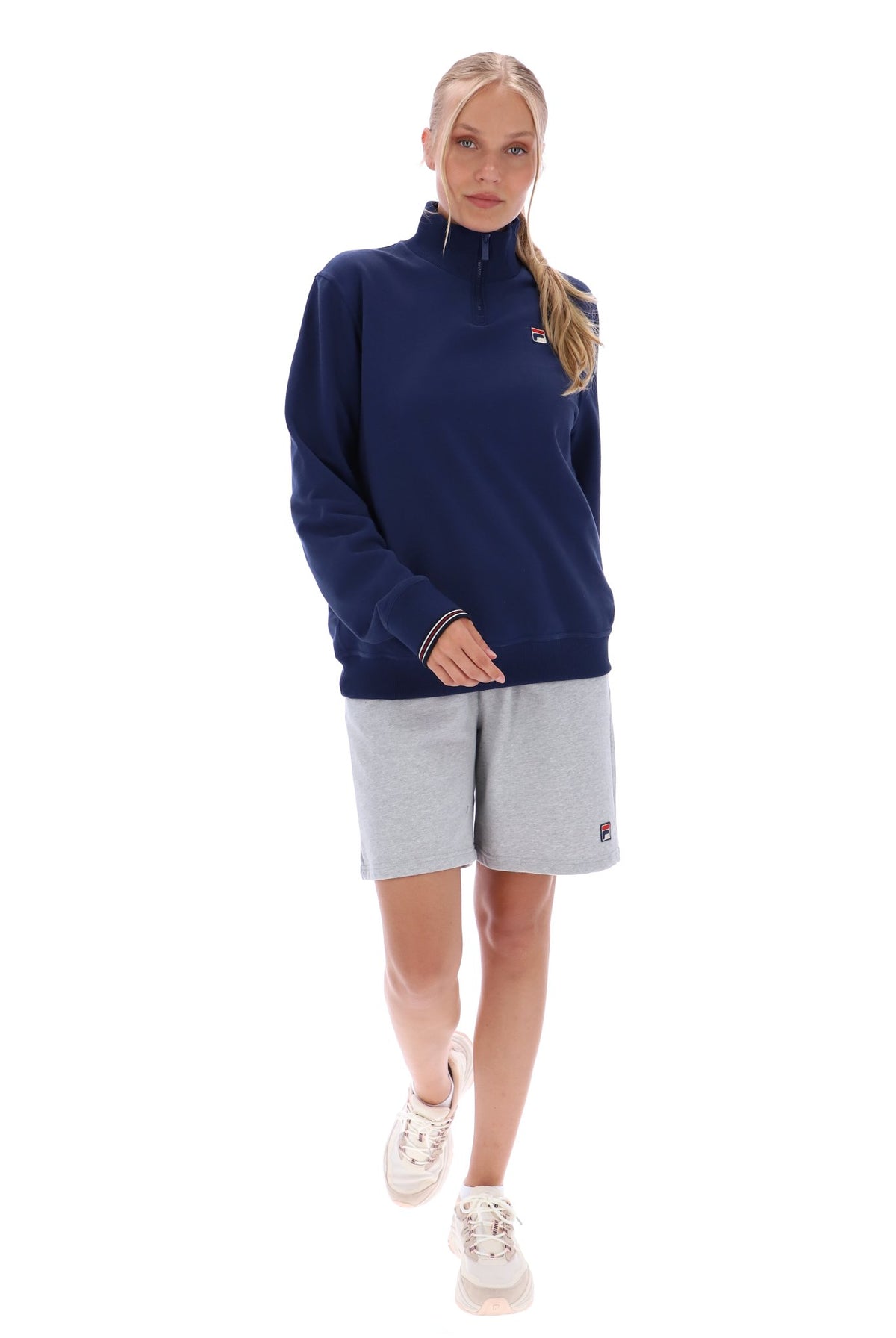 Woman wearing a navy quarter zip jumper with a small FILA logo on the chest, paired with grey FILA jogger shorts, against a white background