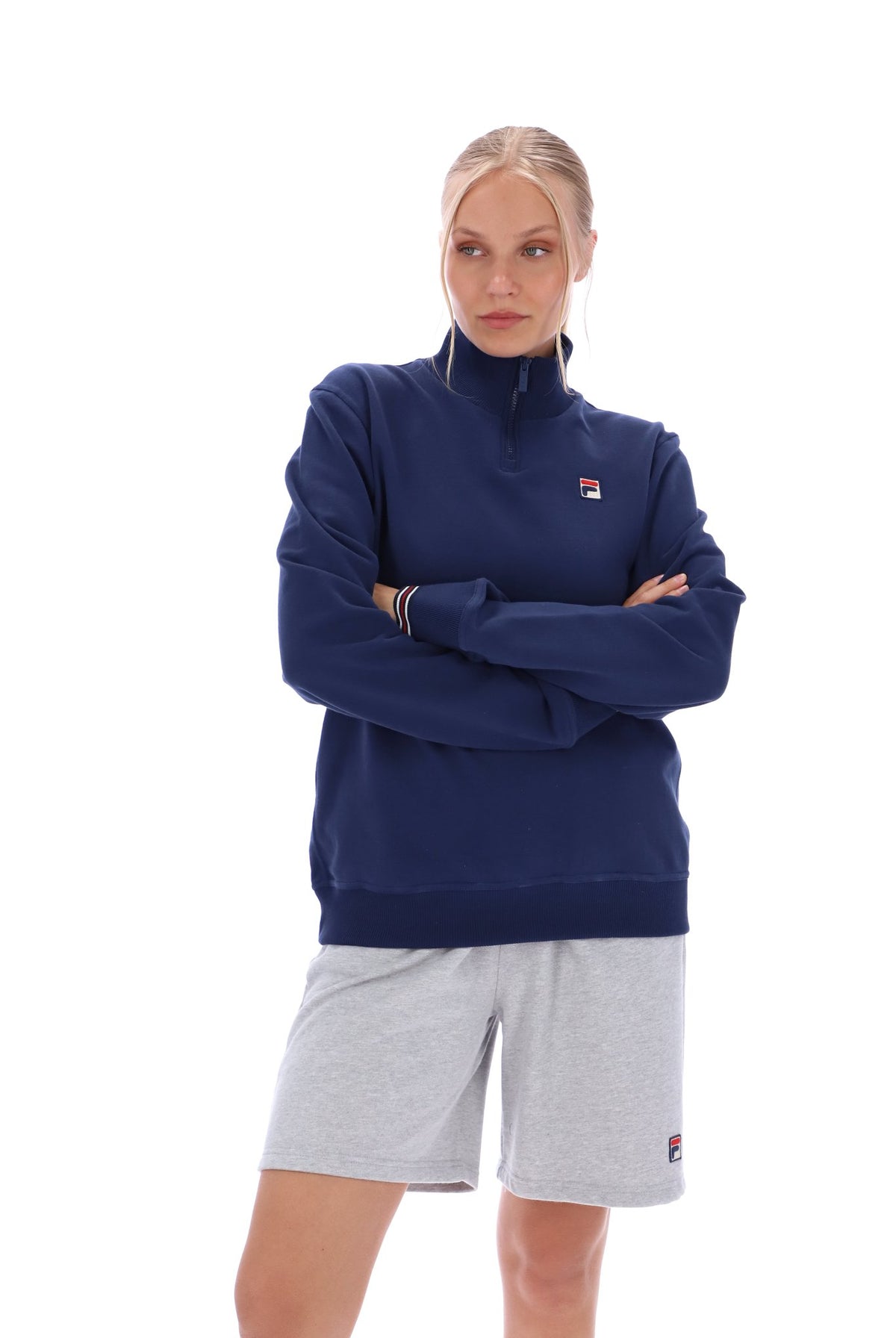Woman wearing a navy quarter zip jumper with a small FILA logo on the chest, paired with grey FILA jogger shorts, against a white background
