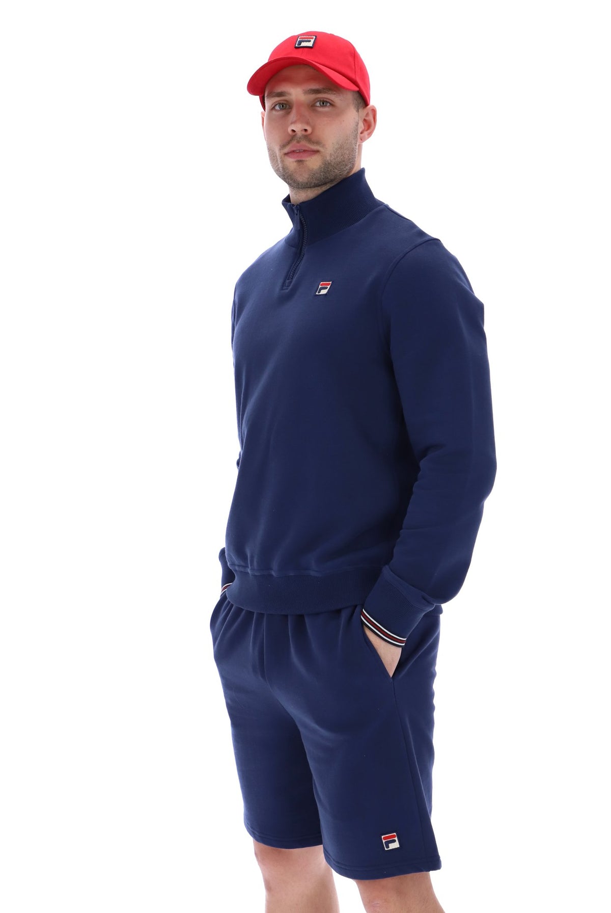 Man wearing a navy quarter zip jumper with a small FILA logo on the chest, paired with grey FILA jogger shorts, against a white background