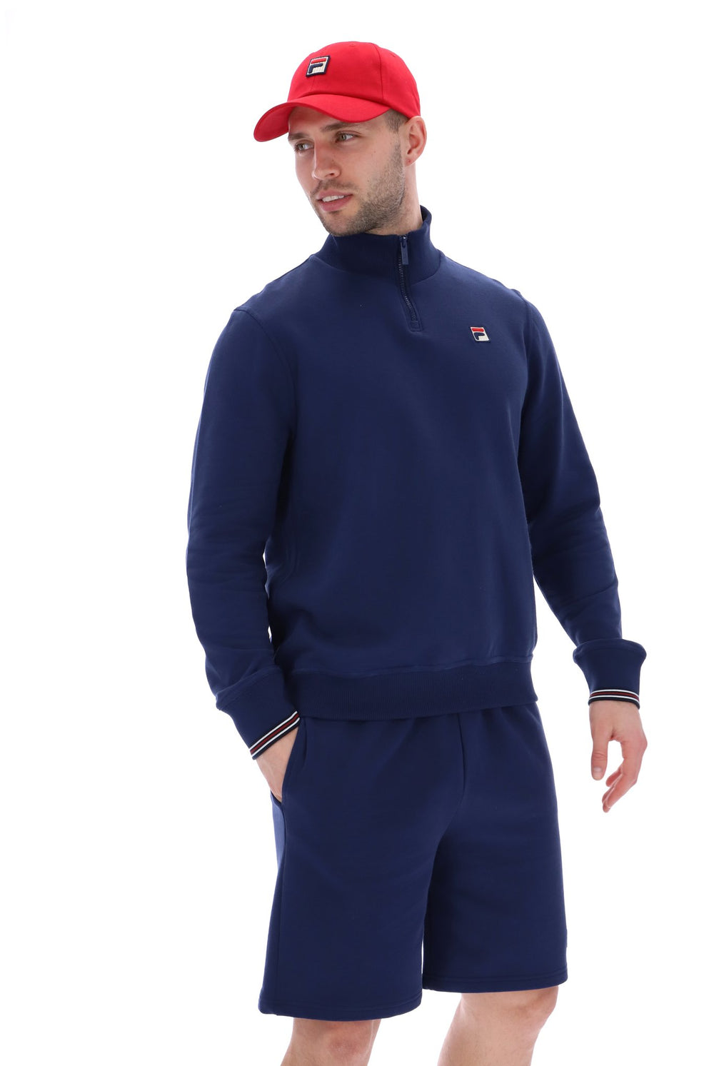 Man wearing a navy quarter zip jumper with a small FILA logo on the chest, paired with grey FILA jogger shorts, against a white background