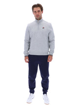 A man modeling a light gray heathered quarter-zip sweatshirt with a small red and white Fila logo on the chest, paired with navy joggers and white sneakers, against a plain white background.