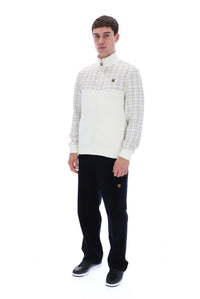 A man modeling a cream and beige plaid quarter-zip pullover sweater with black trousers and black sneakers, standing against a solid white background.