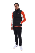 A man modeling a black puffer vest with orange and white color-blocked sleeves, paired with black track pants and white sneakers, on a solid white background.