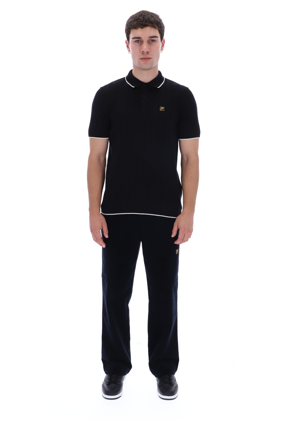 A man modeling a black short-sleeve polo shirt with white piping and a small gold embroidered logo on the chest, paired with black trousers and black sneakers, against a plain white background.
