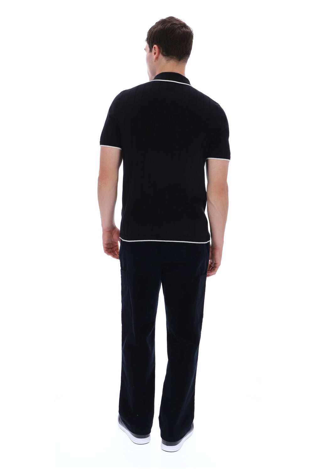 A man viewed from behind wearing a black short-sleeve polo shirt with white piping trim on the collar and sleeve cuffs, paired with dark trousers and white sneakers, against a plain white background.
