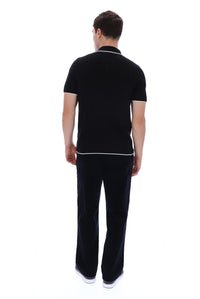 A man viewed from behind wearing a black short-sleeve polo shirt with white piping trim on the collar and sleeve cuffs, paired with dark trousers and white sneakers, against a plain white background.