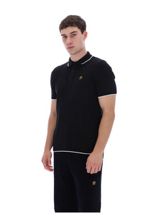 A man modeling a black short-sleeve polo shirt with white piping and a small gold embroidered logo on the chest, paired with matching black trousers, against a plain white background.