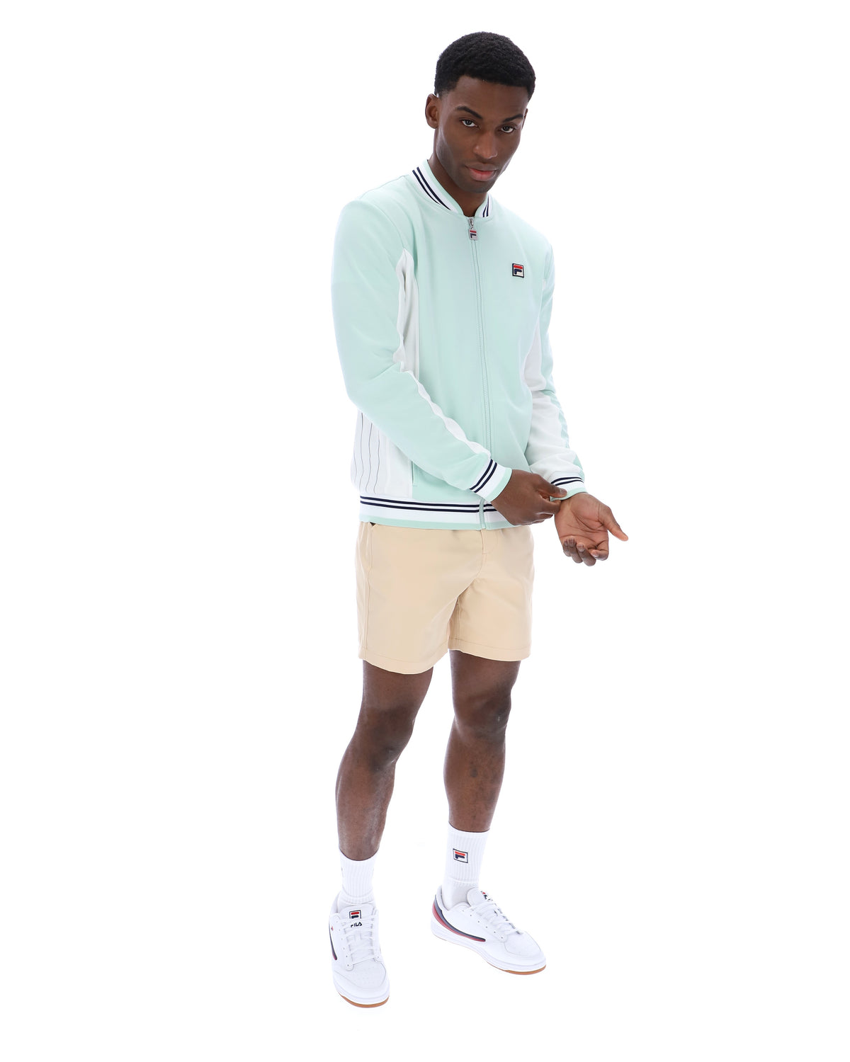 Man wearing a light green jacket with white accents, beige shorts, and white sneakers on a white background