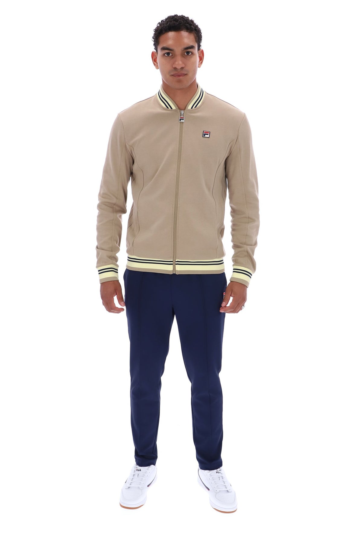 A man modeling a beige Fila zip-up bomber jacket with green and white striped ribbed collar, cuffs, and hem, paired with navy pants and white sneakers, on a solid white background.