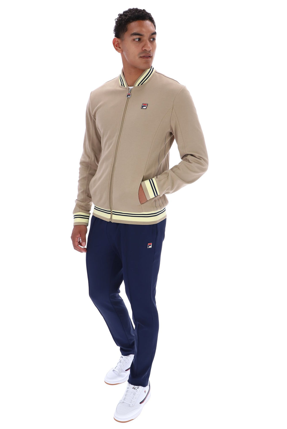 A man modeling a beige zip-up bomber jacket and navy blue sweatpants set by Filippa De Silva, featuring striped ribbed trim and the brand's logo on the chest and thigh.