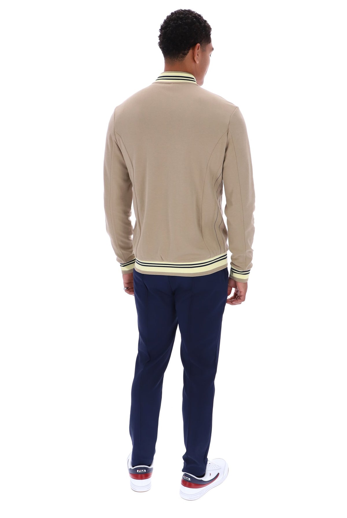 A man viewed from behind wearing a beige long-sleeve knit sweater with green and white striped ribbed collar, cuffs, and hem, paired with navy pants and white sneakers with red accents, on a white background.