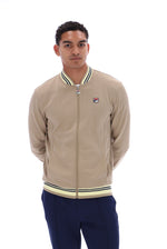 A man wearing a beige Fila zip-up bomber jacket with navy and cream ribbed trim, paired with navy pants, against a solid white background.