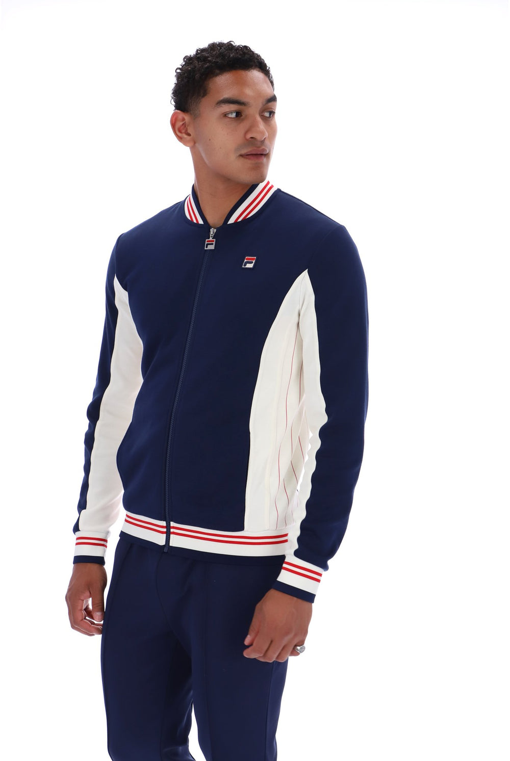 A man wearing a navy blue and white FILA zip-up track jacket with red and white striped ribbed collar, cuffs, and hem, paired with navy pants, against a plain white background.