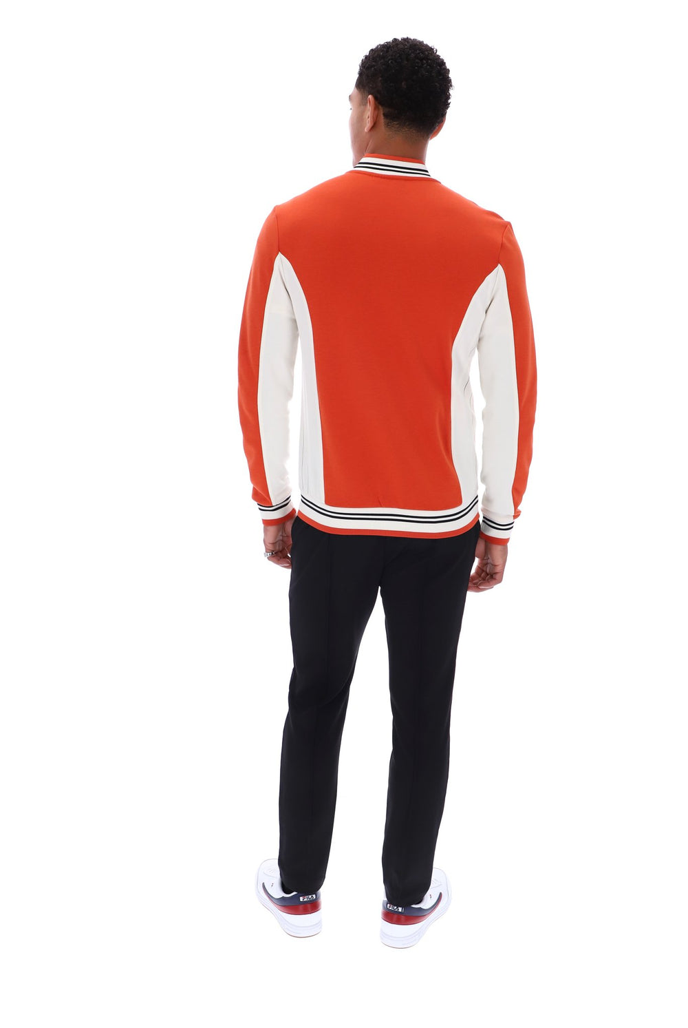 A man viewed from behind wearing a bright orange and white color-blocked knit sweater, black pants, and white sneakers with red accents, set against a plain white background.
