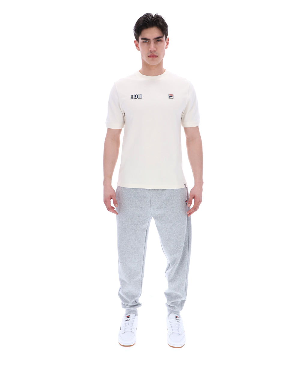 Man wearing a white t-shirt with a FILA logo on the left side of the chest, and 'EST 1911' text on the right side of the chest. Paired with grey joggers, against a white background