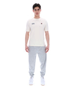 Man wearing a white t-shirt with a FILA logo on the left side of the chest, and 'EST 1911' text on the right side of the chest. Paired with grey joggers, against a white background
