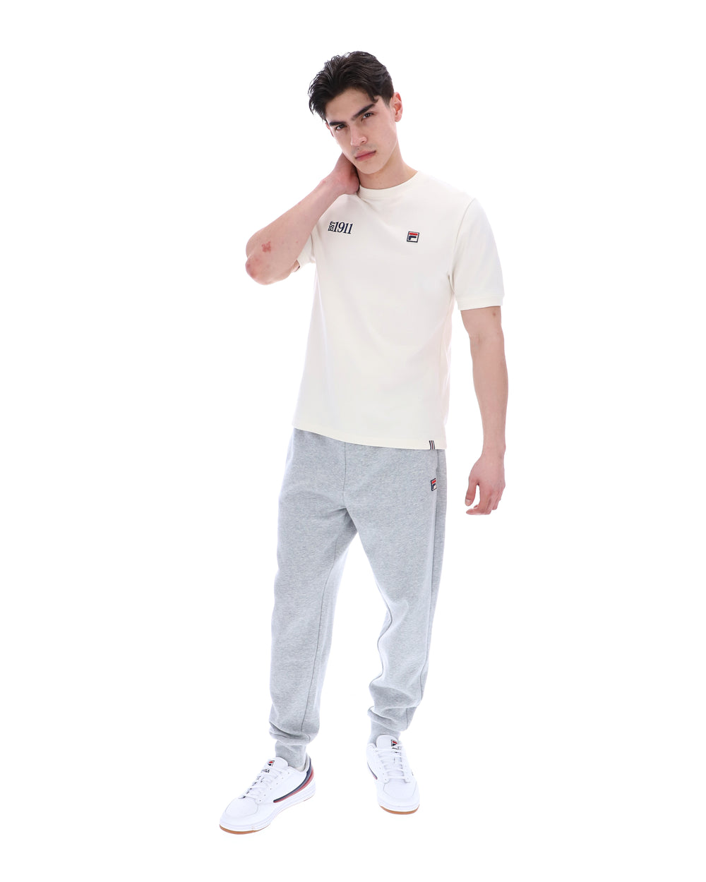 Man wearing a white t-shirt with a FILA logo on the left side of the chest, and 'EST 1911' text on the right side of the chest. Paired with grey joggers, against a white background