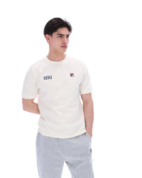 Man wearing a white t-shirt with a FILA logo on the left side of the chest, and 'EST 1911' text on the right side of the chest. Paired with grey joggers, against a white background