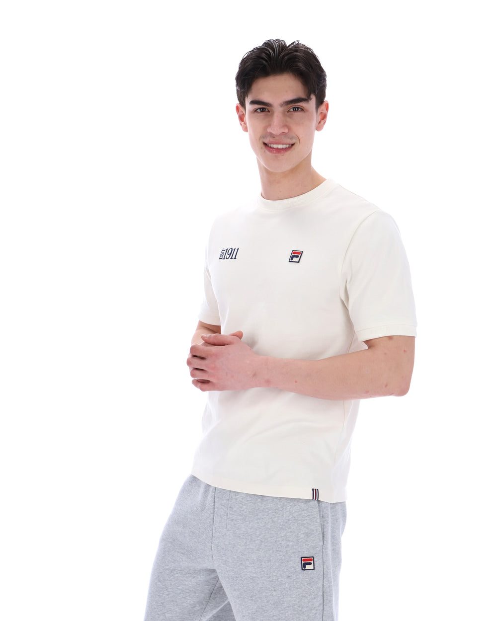 Man wearing a white t-shirt with a FILA logo on the left side of the chest, and 'EST 1911' text on the right side of the chest. Paired with grey joggers, against a white background