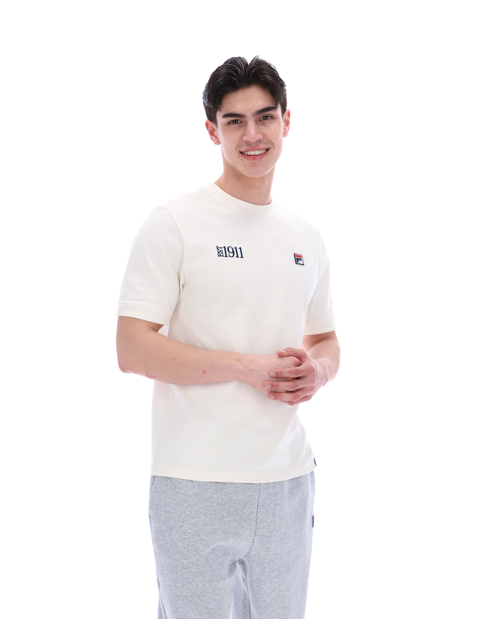 Man wearing a white t-shirt with a FILA logo on the left side of the chest, and 'EST 1911' text on the right side of the chest. Paired with grey joggers, against a white background