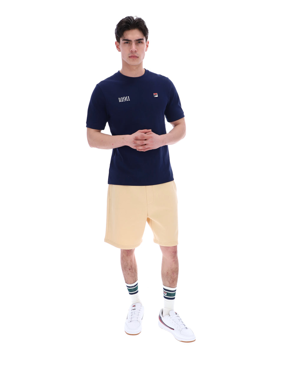 Man wearing a navy blue t-shirt with a FILA logo on the left side of the chest, and 'EST 1911' text on the right side of the chest. Paired with a beige shorts, against a white background