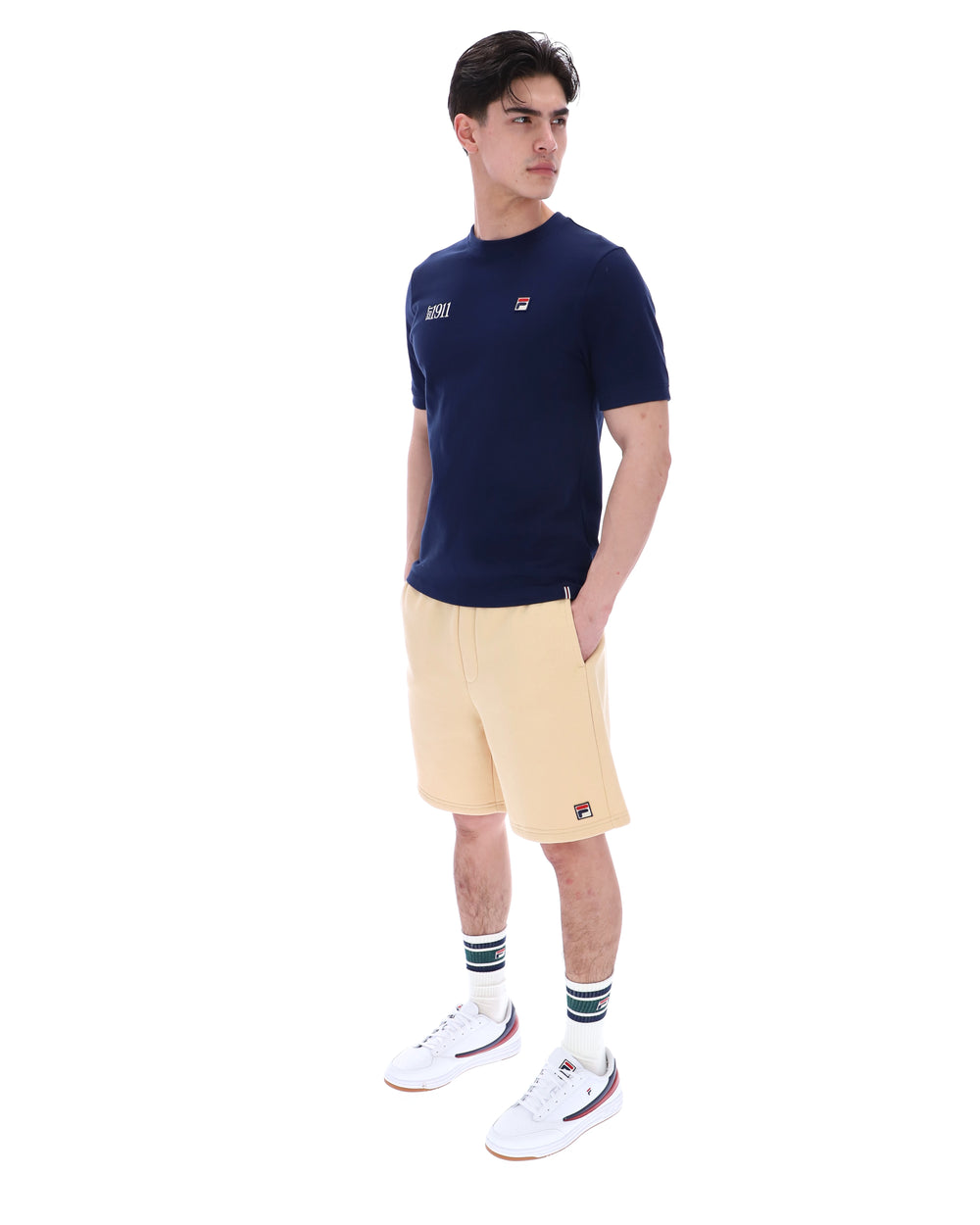 Man wearing a navy blue t-shirt with a FILA logo on the left side of the chest, and 'EST 1911' text on the right side of the chest. Paired with a beige shorts, against a white background