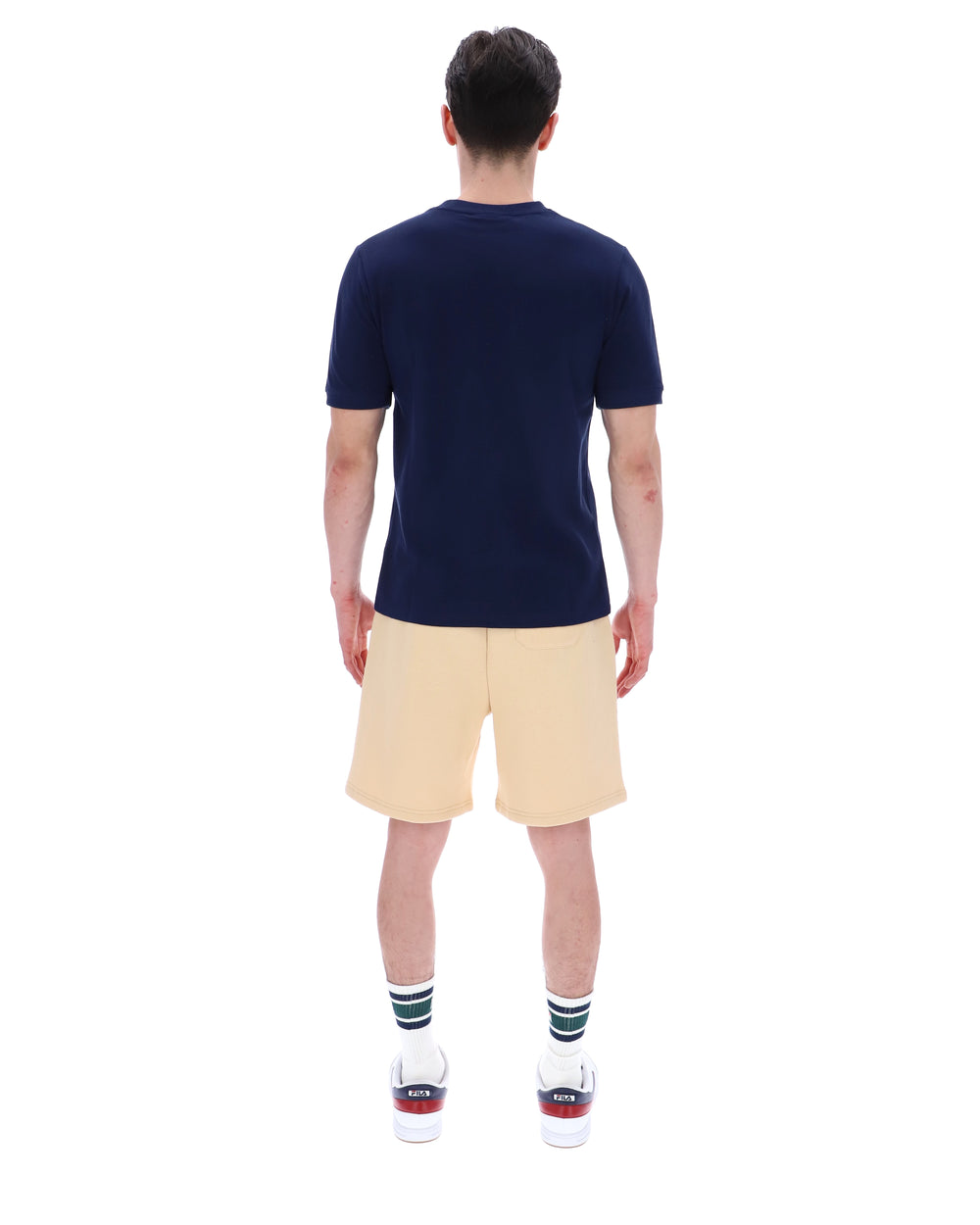 Man wearing a navy blue t-shirt with a FILA logo on the left side of the chest, and 'EST 1911' text on the right side of the chest. Paired with a beige shorts, against a white background