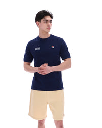 Man wearing a navy blue t-shirt with a FILA logo on the left side of the chest, and 'EST 1911' text on the right side of the chest. Paired with a beige shorts, against a white background