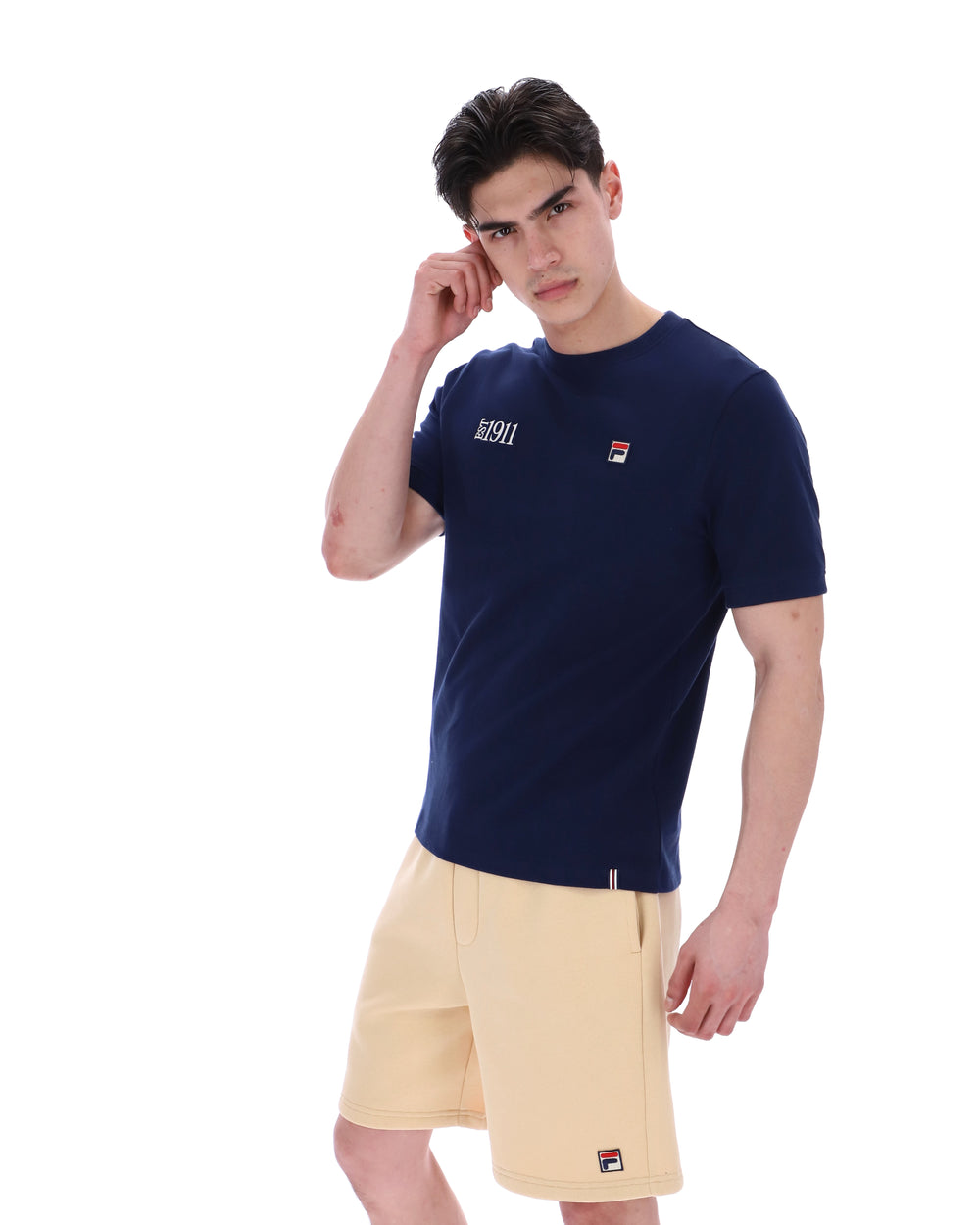 Man wearing a navy blue t-shirt with a FILA logo on the left side of the chest, and 'EST 1911' text on the right side of the chest. Paired with a beige shorts, against a white background