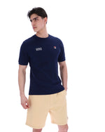 Man wearing a navy blue t-shirt with a FILA logo on the left side of the chest, and 'EST 1911' text on the right side of the chest. Paired with a beige shorts, against a white background