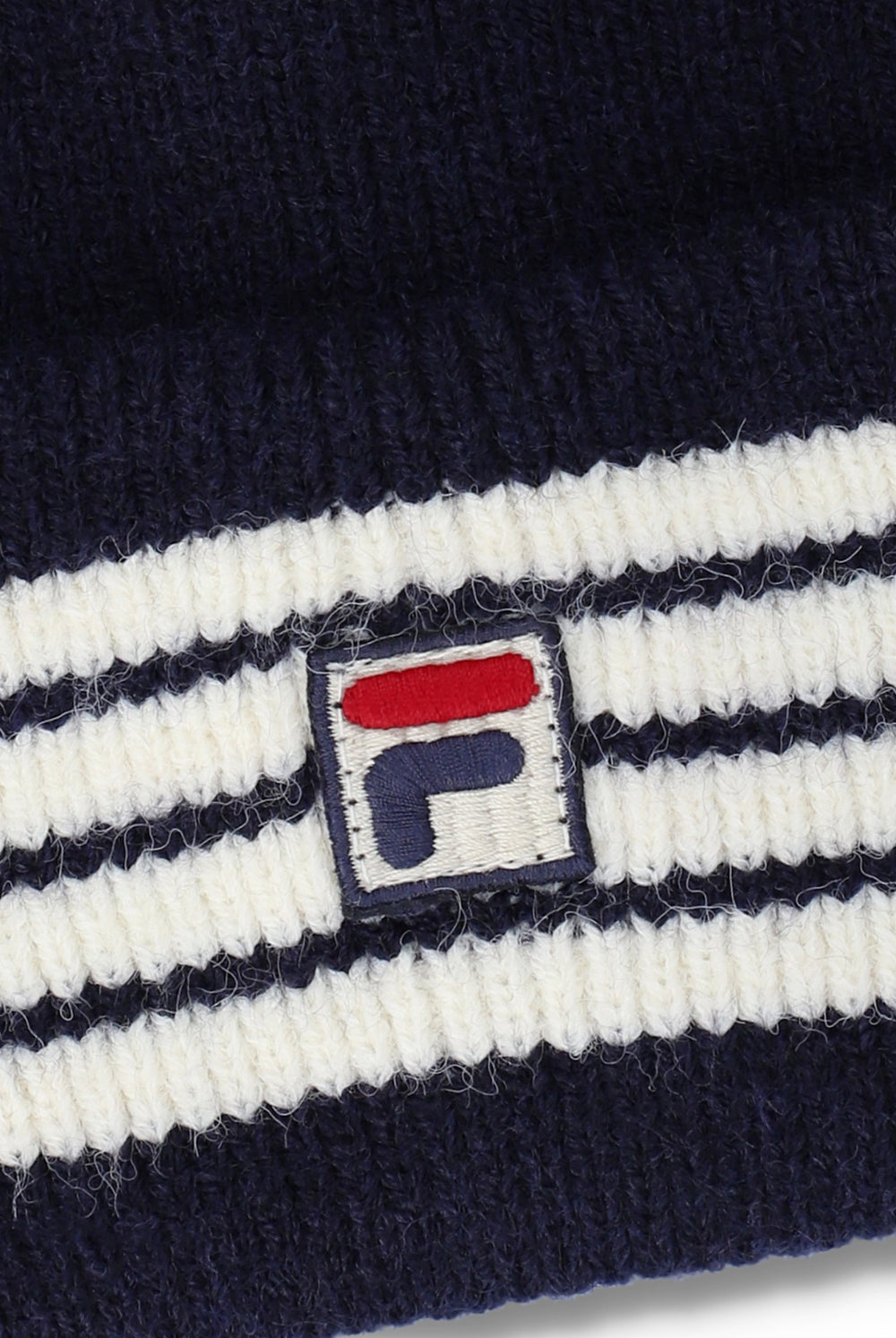 Close-up of a ribbed knit beanie in navy blue and white stripes, featuring a small embroidered Fila logo patch on the front.