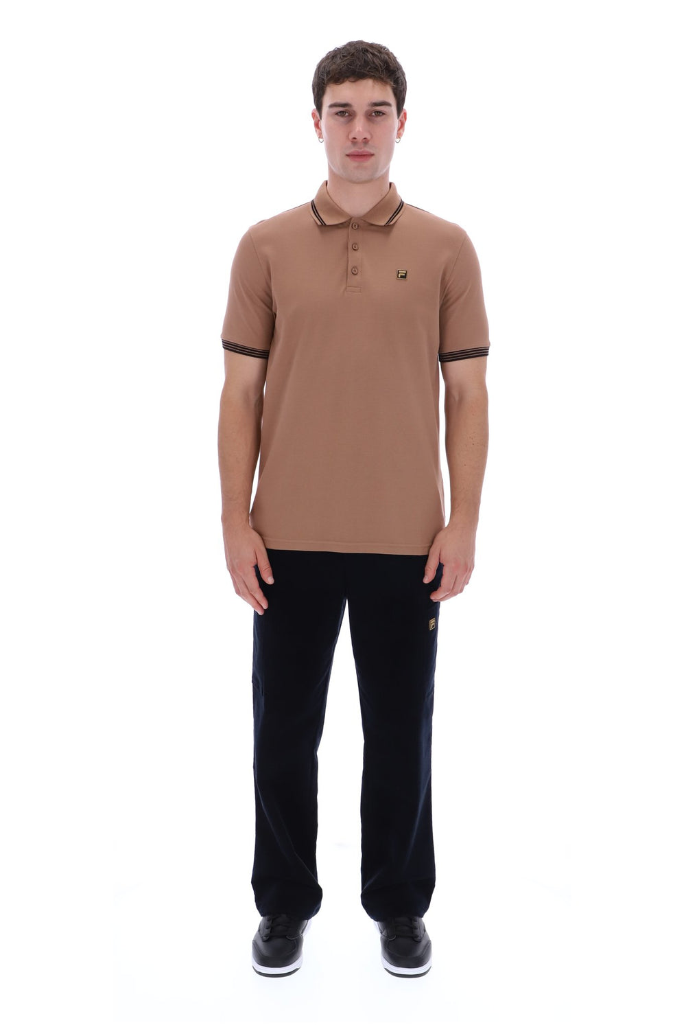 A man modeling a tan short-sleeve polo shirt with dark trim on the collar and sleeve cuffs, paired with black trousers and black sneakers, against a plain white background.