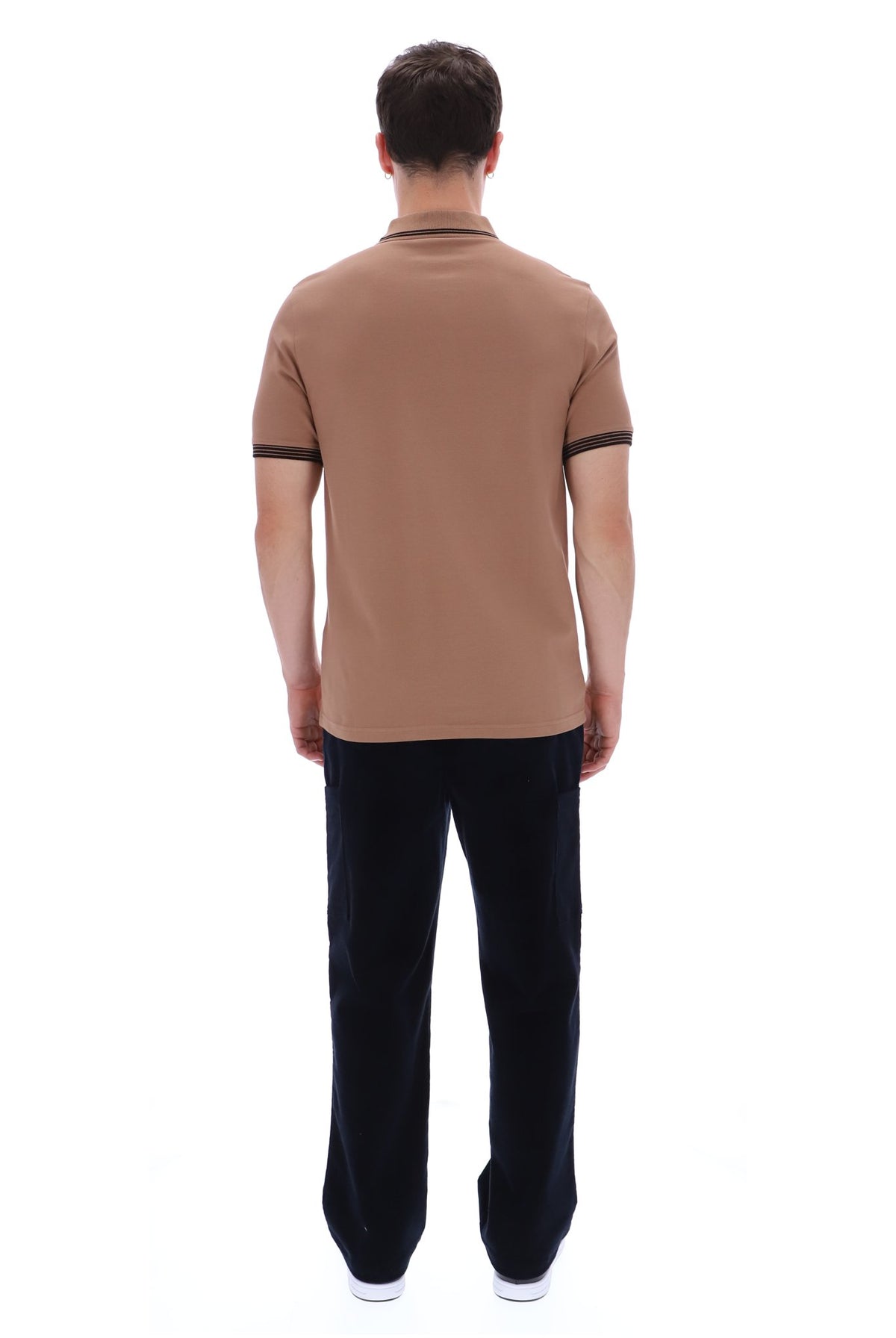 A man viewed from behind wearing a tan short-sleeve polo shirt with black trim on the collar and sleeve cuffs, paired with black trousers and white sneakers, against a plain white background.