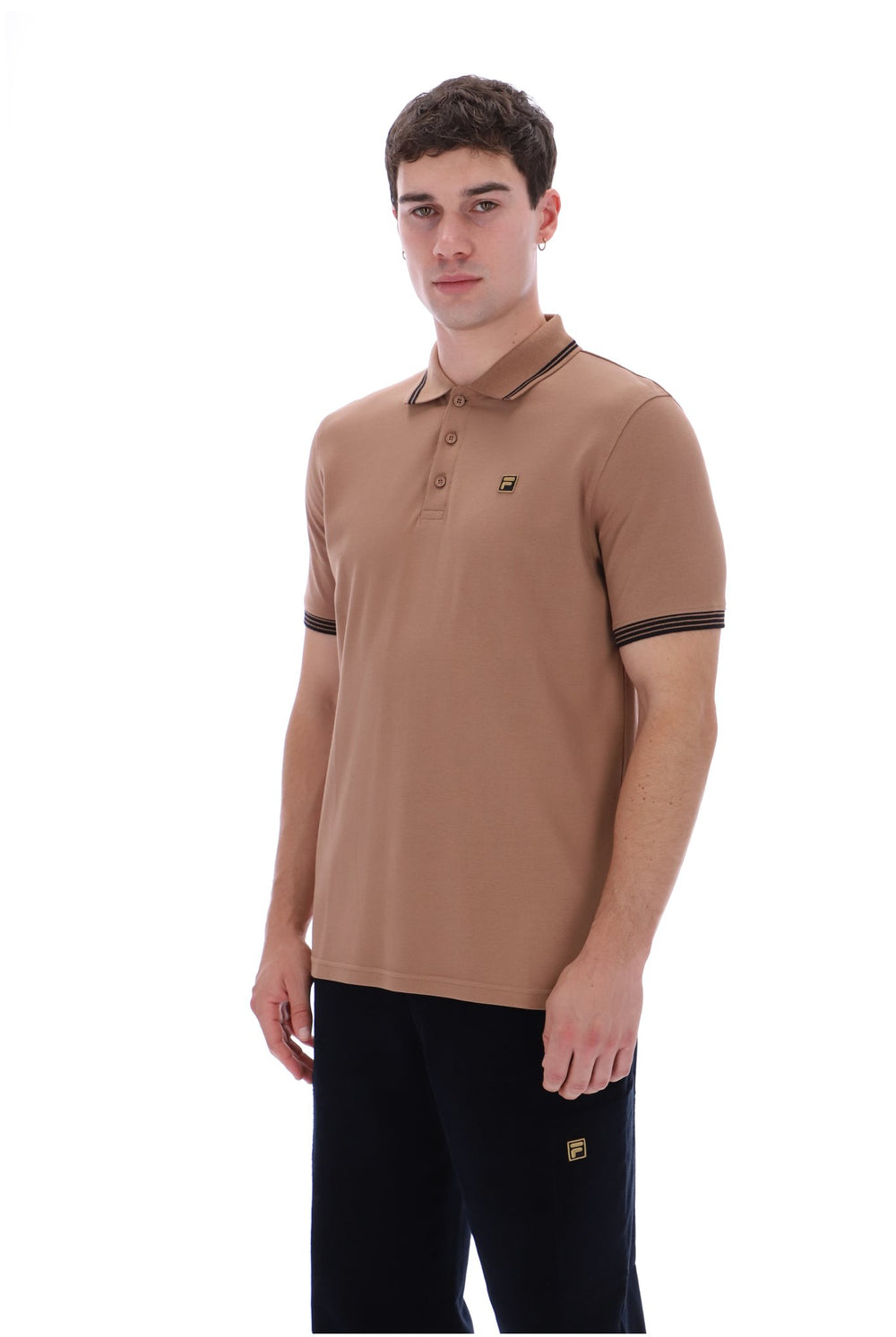 A man wearing a tan short-sleeve polo shirt with dark trim on the collar, sleeve cuffs, and a small embroidered logo on the chest, paired with black pants, against a plain white background.