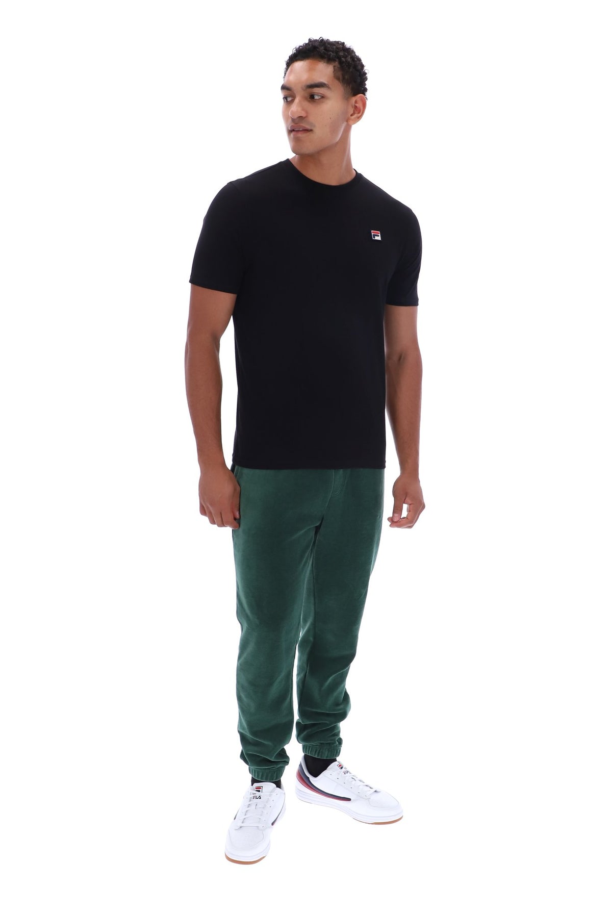A man modeling a black short-sleeve crewneck t-shirt with a small red and white logo on the left chest, paired with dark green jogger pants and white sneakers, shot against a solid white background.