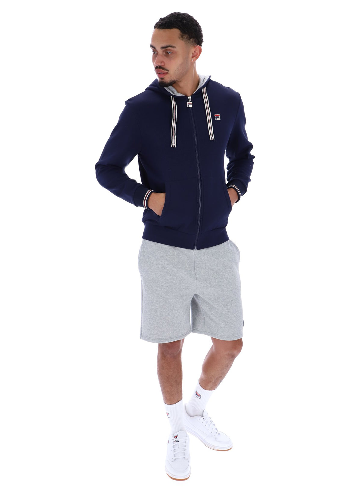 A male model wearing a navy blue zip-up hoodie with the FILA logo on the chest, paired with light grey shorts.