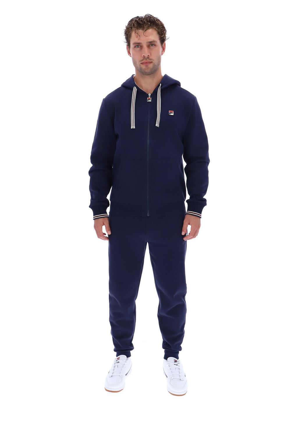 A man modeling a navy blue zip-up hoodie and matching sweatpants set with white drawstrings and white sneakers, photographed against a solid white background.