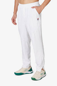 A person wearing white tennis court track pants with the FILA logo on the left leg and white sneakers.