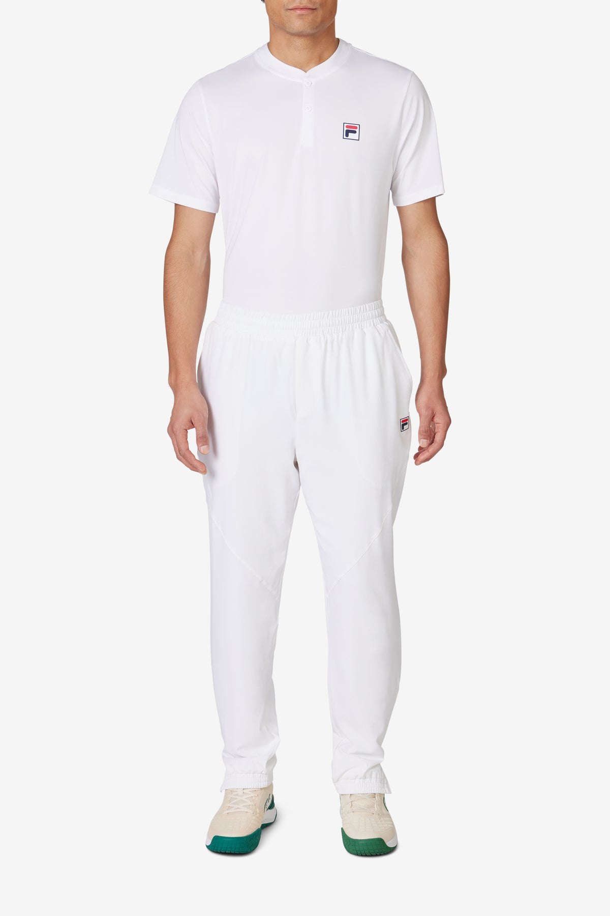 A person wearing white tennis court track pants with the FILA logo on the left leg and white sneakers.