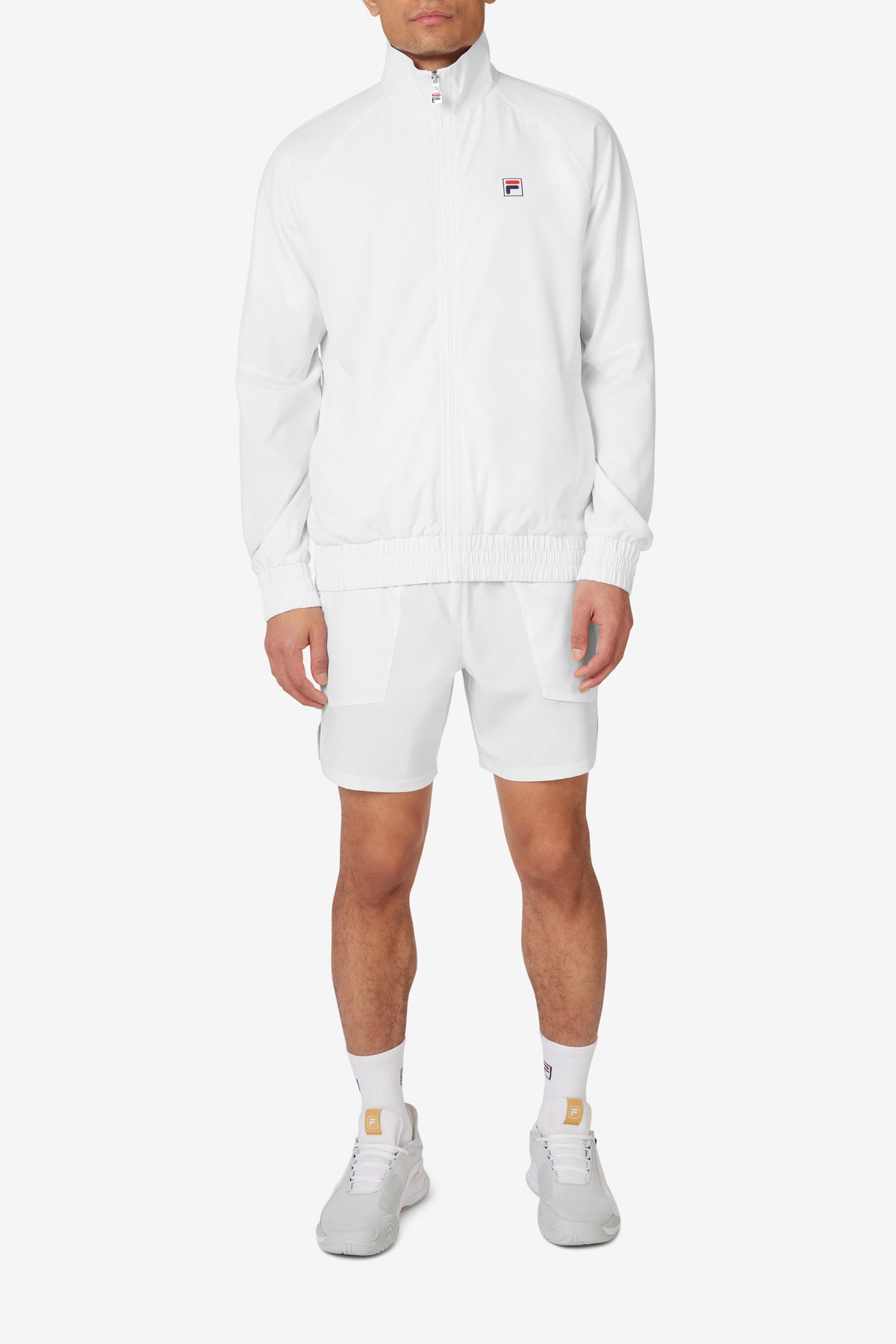 A man wearing a white FILA tennis court track jacket with a zipper, collar, and matching white shorts.