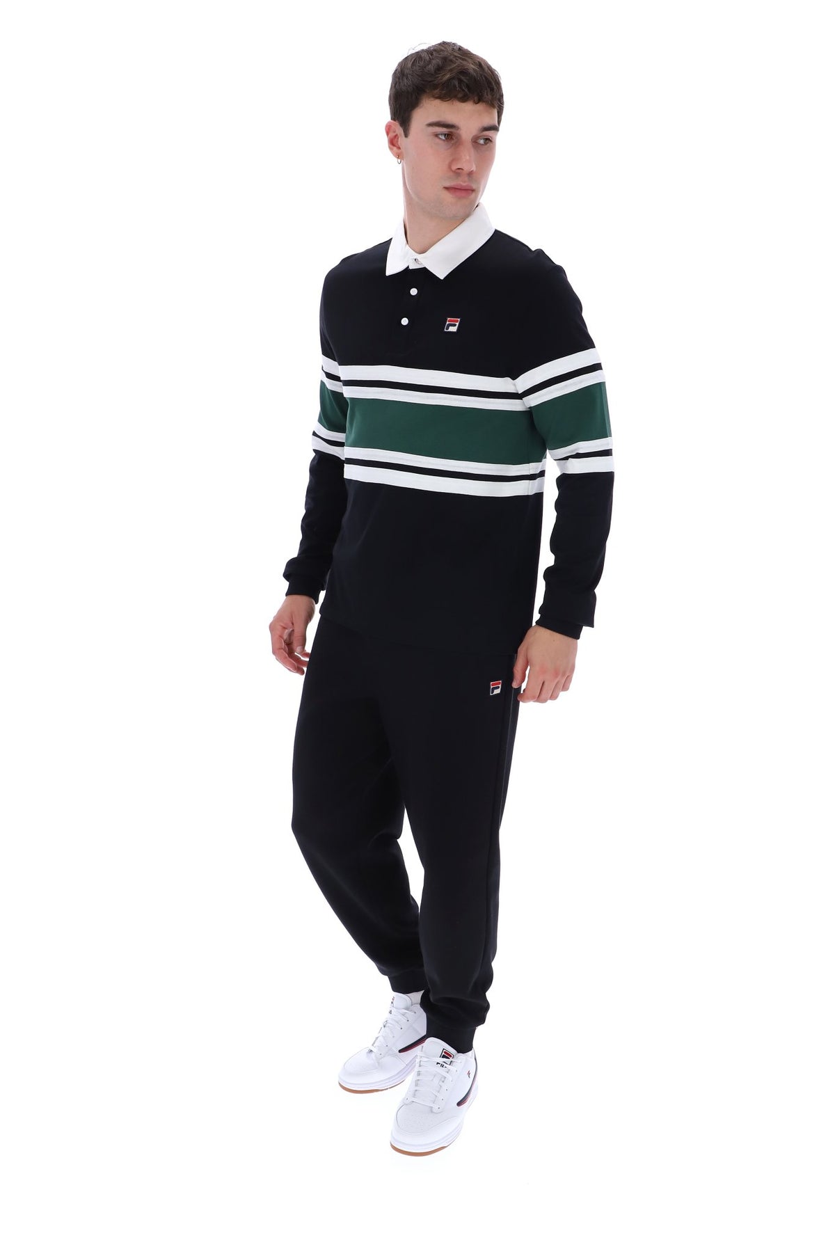 A young man modeling a black long-sleeve rugby-style polo shirt with green and white horizontal stripes on the chest and sleeves, paired with black joggers and white sneakers, against a plain white background.
