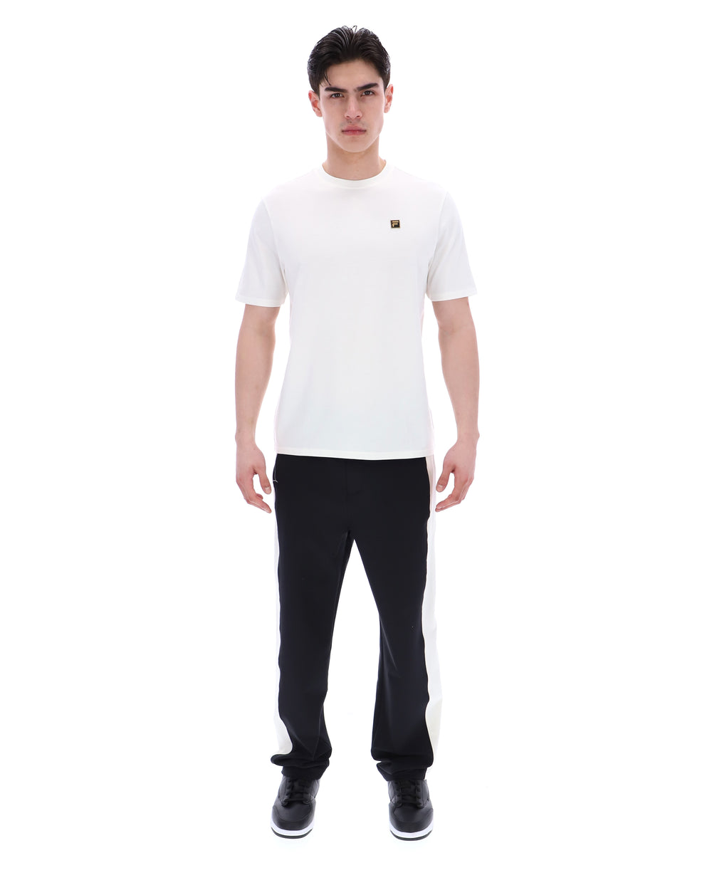 Man wearing a white t-shirt with a small FILA gold logo, paired with black pants, against a white background