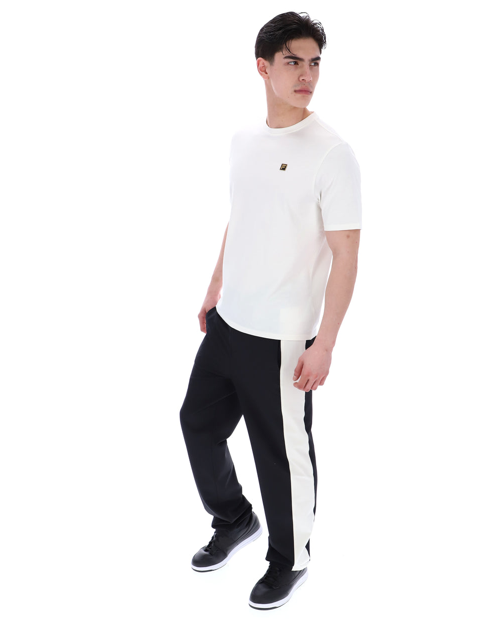Man wearing a white t-shirt with a small FILA gold logo, paired with black pants, against a white background