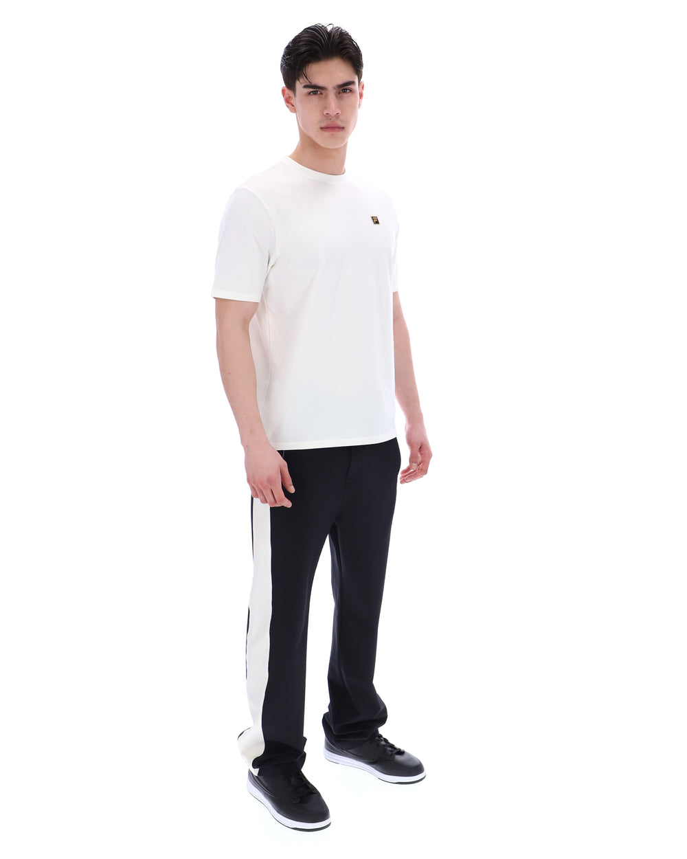 Man wearing a white t-shirt with a small FILA gold logo, paired with black pants, against a white background