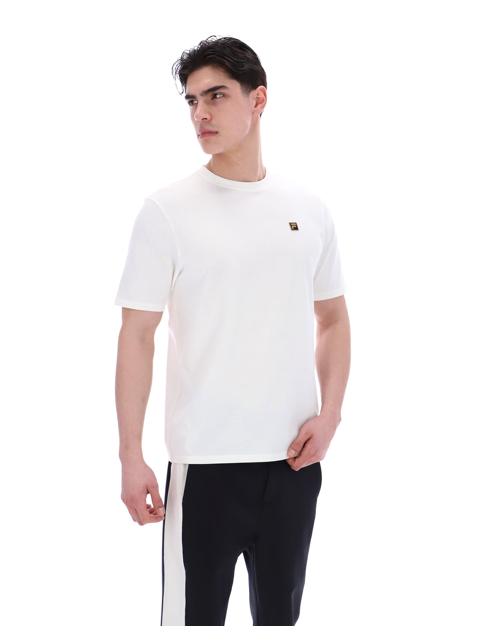 Man wearing a white t-shirt with a small FILA gold logo, paired with black pants, against a white background