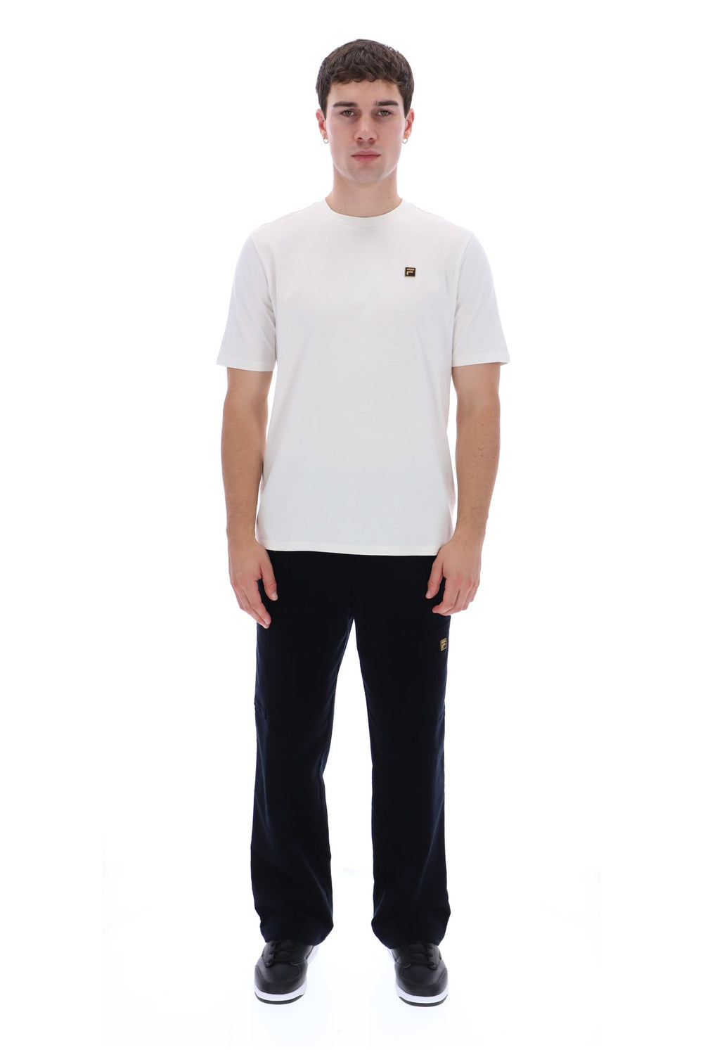 A man modeling a white short-sleeve crewneck t-shirt with a small brown embroidered logo on the left chest, paired with black sweatpants and black sneakers, against a solid white background.