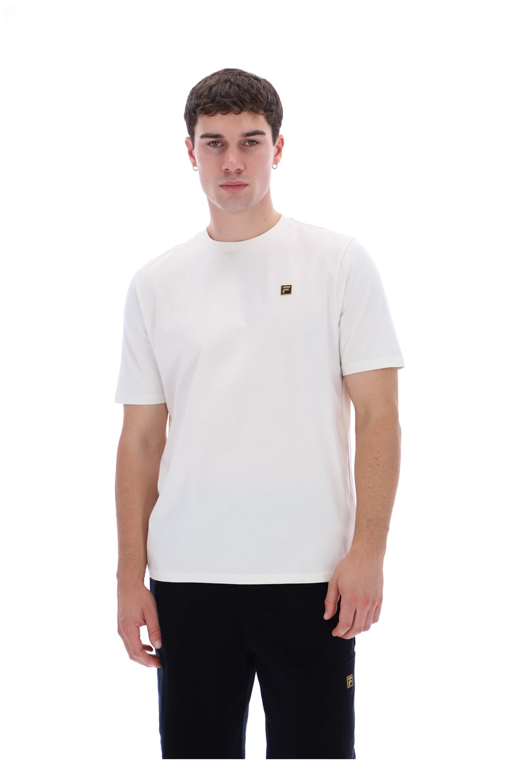 A man wearing a white crew-neck t-shirt with a small gold square logo on the left chest, paired with black pants, against a solid white background.