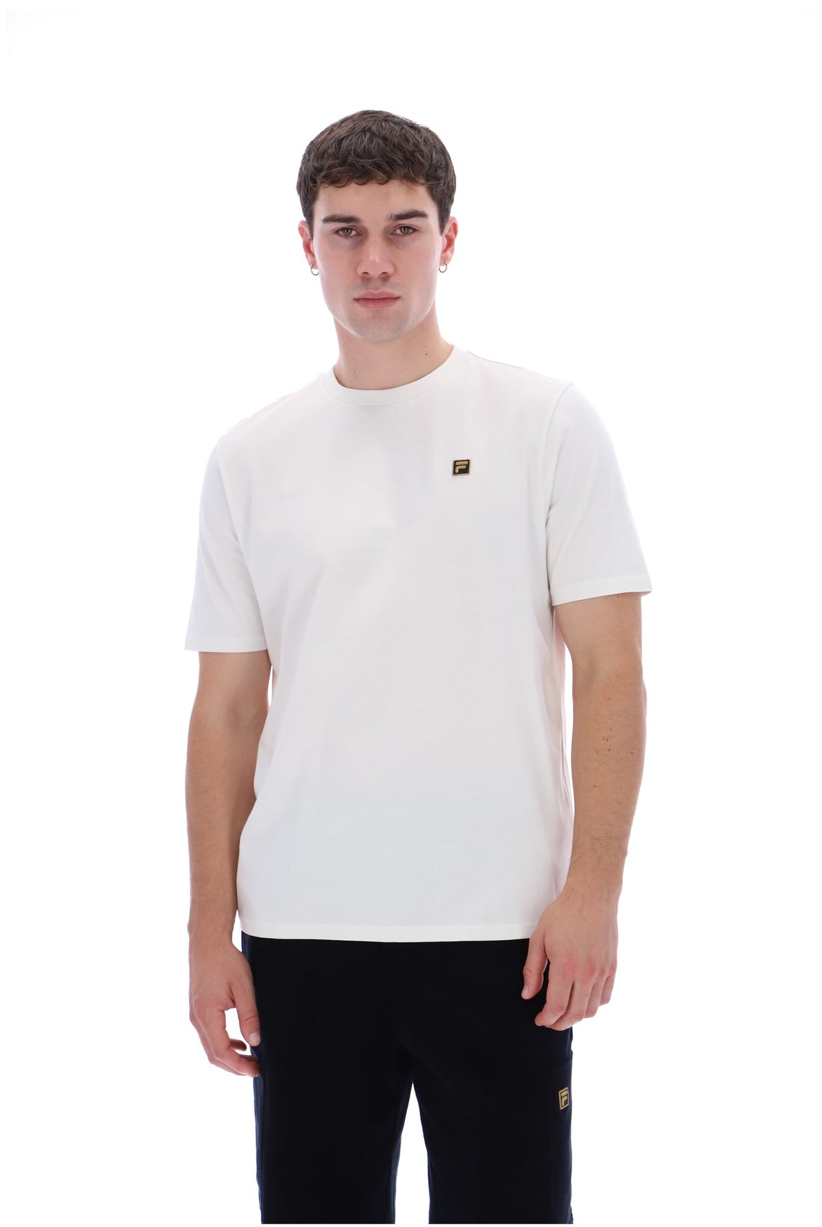 A man wearing a white crew-neck t-shirt with a small gold square logo on the left chest, paired with black pants, against a solid white background.